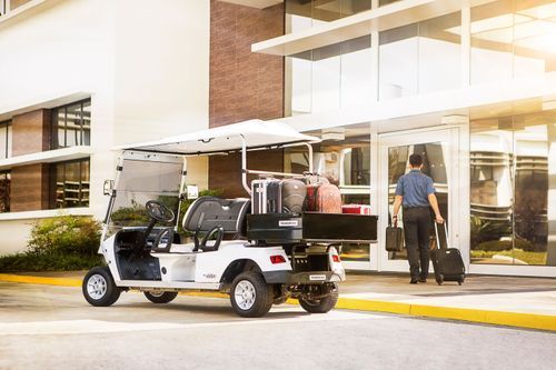 Veículo elétrico utilitário branco Tramontina com caçamba traseira carregada de malas, estacionado na entrada de um hotel enquanto funcionário transporta bagagem.
