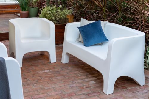 Sofá e poltronas em plástico rígido branco de linhas arredondadas com almofada azul dispostos em pátio externo com piso de tijolo.