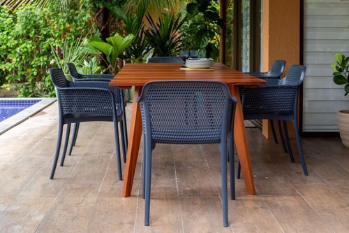 Mesa retangular de madeira natural cercada por cadeiras de plástico perfuradas na cor cinza-escura em uma varanda com piscina ao fundo.