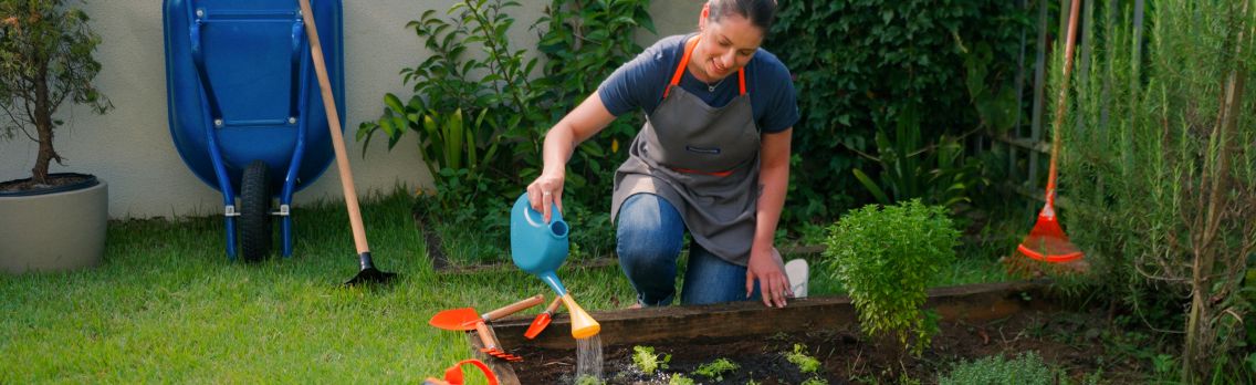Banner horizontal mostrando uma mulher cuidando de uma horta. Ela usa um avental cinza e rega as plantas com um regador azul. Ao fundo, um carrinho de mão azul e ferramentas de jardinagem laranjas sobre o gramado verde.