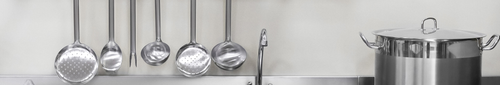Conchas, escumadeiras e garfo trinchante em aço inox pendurados em suporte de parede ao lado de caldeirão industrial com tampa em cozinha profissional.