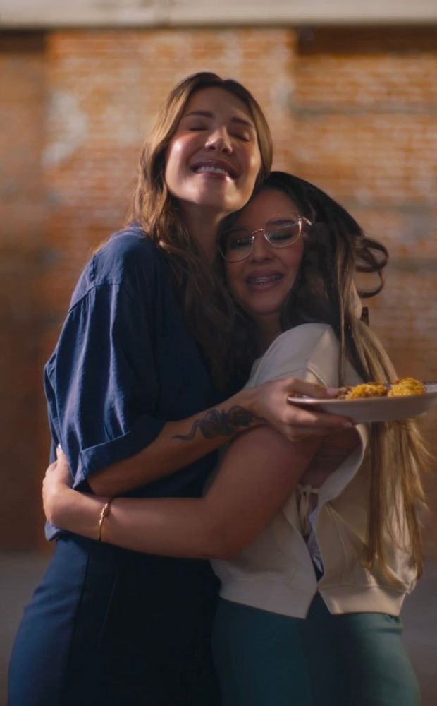 Fotografia de duas mulheres se abraçando afetuosamente. A mulher à esquerda tem cabelos castanhos longos, veste uma blusa azul-escura, tem uma tatuagem no antebraço e sorri com os olhos fechados. A mulher à direita tem cabelos longos, usa óculos de grau, 