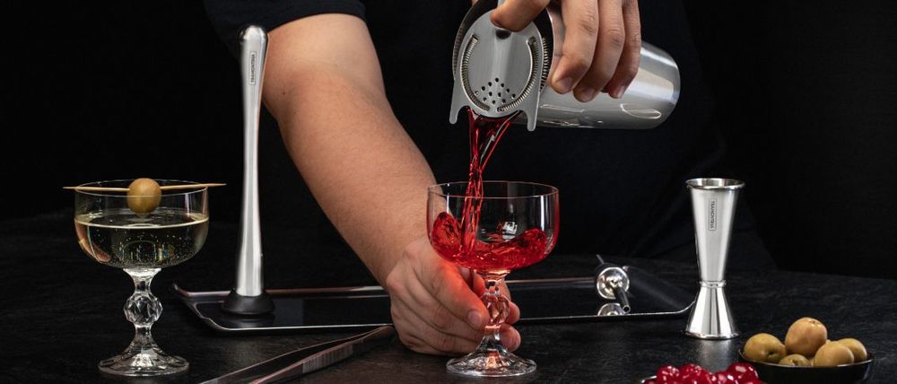 Conjunto de utensílios de bar em inox, com coqueteleira, dosador e colher de bar, sendo utilizado para preparar drinks em taças elegantes. Estilo sofisticado para coquetéis.