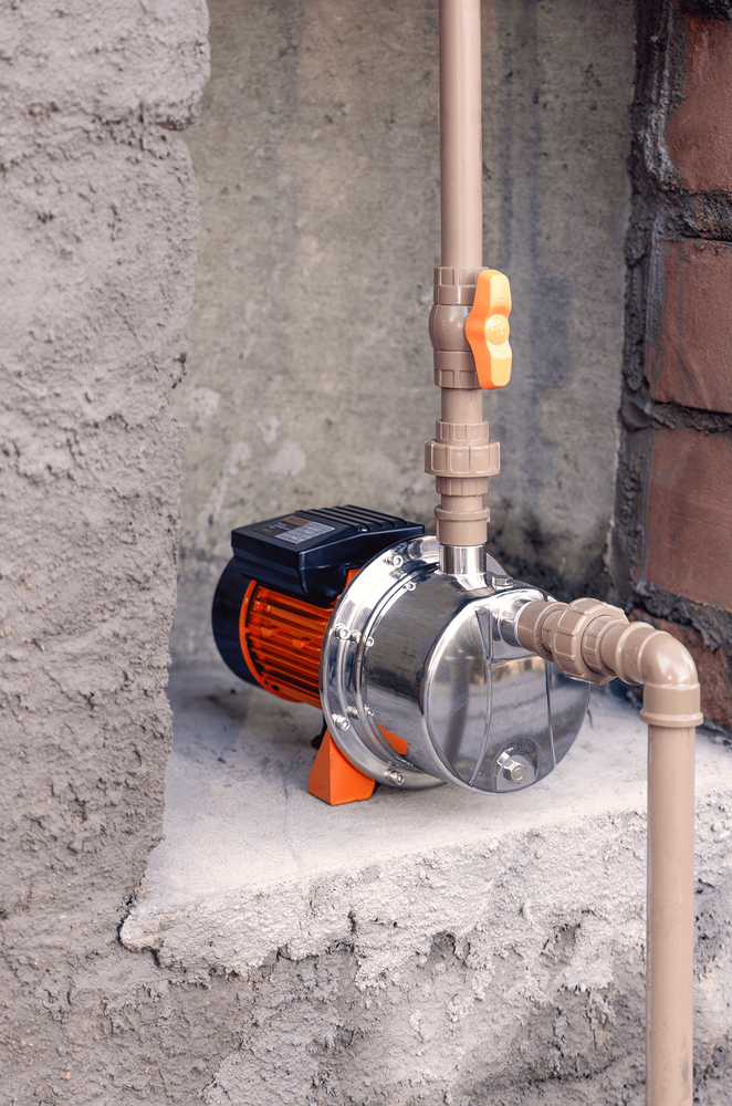 Uma eletrobomba de água com corpo em aço inoxidável e motor laranja instalada sobre uma base de concreto. A bomba está conectada a canos de PVC beges, com uma válvula de fechamento laranja visível na tubulação vertical.