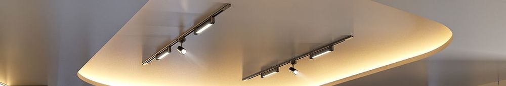 Vista aproximada de um sistema de iluminação de trilho Tramontina em metal preto com cinco spots direcionáveis acesos, instalados em um teto de concreto aparente com estilo industrial.