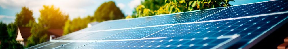 Vista em close de painéis de energia solar instalados no telhado de uma casa, refletindo a luz do sol com árvores e céu azul ao fundo em um dia ensolarado.