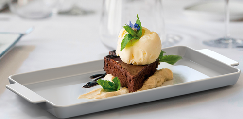 Prato travessa retangular de porcelana Tramontina servido com fatia de brownie, bola de sorvete de creme e calda em mesa posta com taças ao fundo.