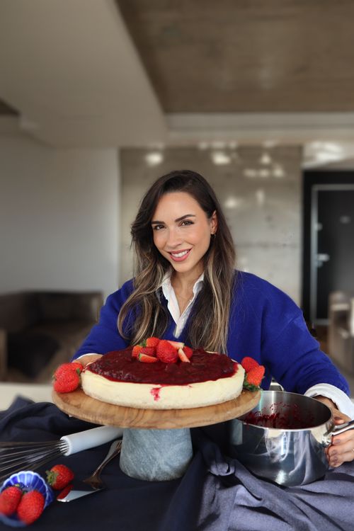 Mulher sorrindo, segurando um cheesecake de morangos em uma tábua de madeira enquanto, com a outra mão, segura uma panela de inox da Tramontina. A imagem reflete o prazer de cozinhar em um ambiente acolhedor.