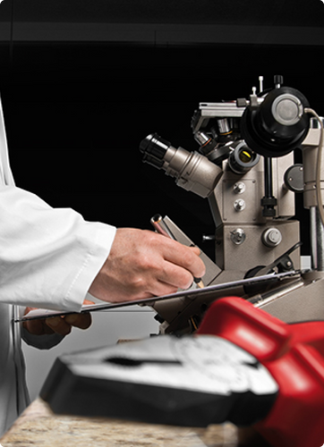 Una persona anotando en un tablero. Al fondo, un microscopio. A frente, la punta de un alicate.
