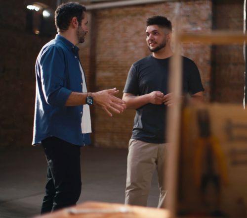 Fotografia de dois homens conversando em um amplo espaço com paredes rústicas de tijolos. À esquerda, Iberê Thenório, vestindo camisa azul aberta sobre uma camiseta branca, gesticula com as mãos de forma explicativa. À direita, um jovem de camiseta escura