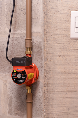 Bomba pressurizadora Tramontina BTCP120 na cor laranja com painel de controle preto, instalada verticalmente em uma tubulação residencial sobre parede de concreto, com a chave seletora nas opções manual, auto e off.