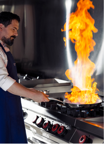 Um chef de perfil em uma cozinha profissional operando uma frigideira com uma grande chama laranja (técnica de flambar) sobre um fogão industrial de aço inox com botões vermelhos.