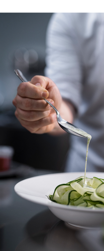 Close-up de uma mão segurando uma colher de metal, despejando um molho branco cremoso com ervas sobre um prato de fitas de abobrinha verde em uma tigela branca.