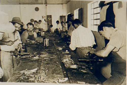 Fotografia antiga mostrando vários artesãos trabalhando em uma longa bancada de madeira na montagem manual de canivetes.