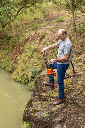 Homem em pé sobre uma rocha à beira de um lago ou rio, segurando uma motobomba submersível Tramontina nas cores laranja e preto, preparando o equipamento com mangueira e cabos elétricos para drenagem ou captação de água.