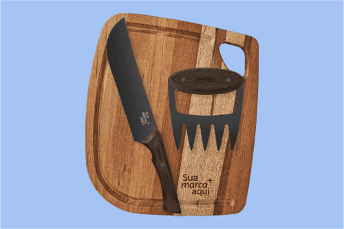 Kit de churrasco Tramontina sobre fundo azul claro. O conjunto contém uma tábua de madeira com canaletas, uma faca de lâmina preta e uma garra de urso para desfiar carne. Tanto a tábua quanto a lâmina da faca exibem a gravação 'Sua marca aqui'. Os cabos s
