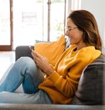 Mulher de óculos e blusa de tricô amarela sorrindo enquanto navega no smartphone sentada confortavelmente em um sofá cinza.