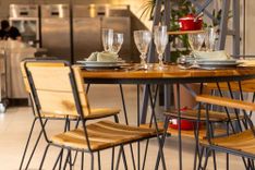 Mesa redonda posta com taças e pratos cercada por cadeiras com assento de madeira e estrutura metálica preta, em um ambiente decorado de restaurante.
