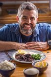 Homem de barba sorridente com uma panela de ferro Tramontina na mesa, repleta de feijoada e acompanhamentos. A imagem retrata um momento de refeição em família, com foco na celebração e na culinária afetiva.