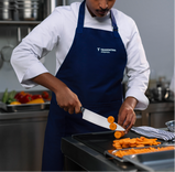 Close-up de um chef usando um avental azul e dólmã branca, cortando cenouras em rodelas com uma faca de chef afiada sobre uma tábua de corte preta em uma cozinha profissional.